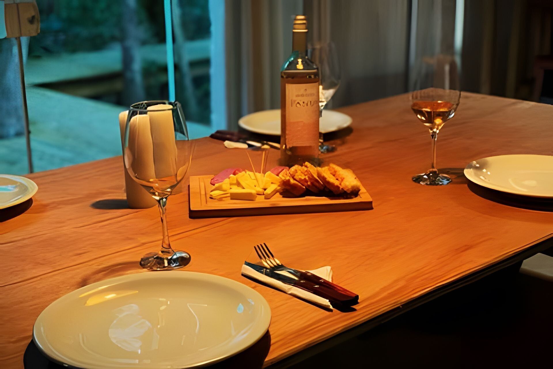 Una mesa de comedor con platos, cubiertos, una copa de vino, una botella y una tabla de madera con comida.