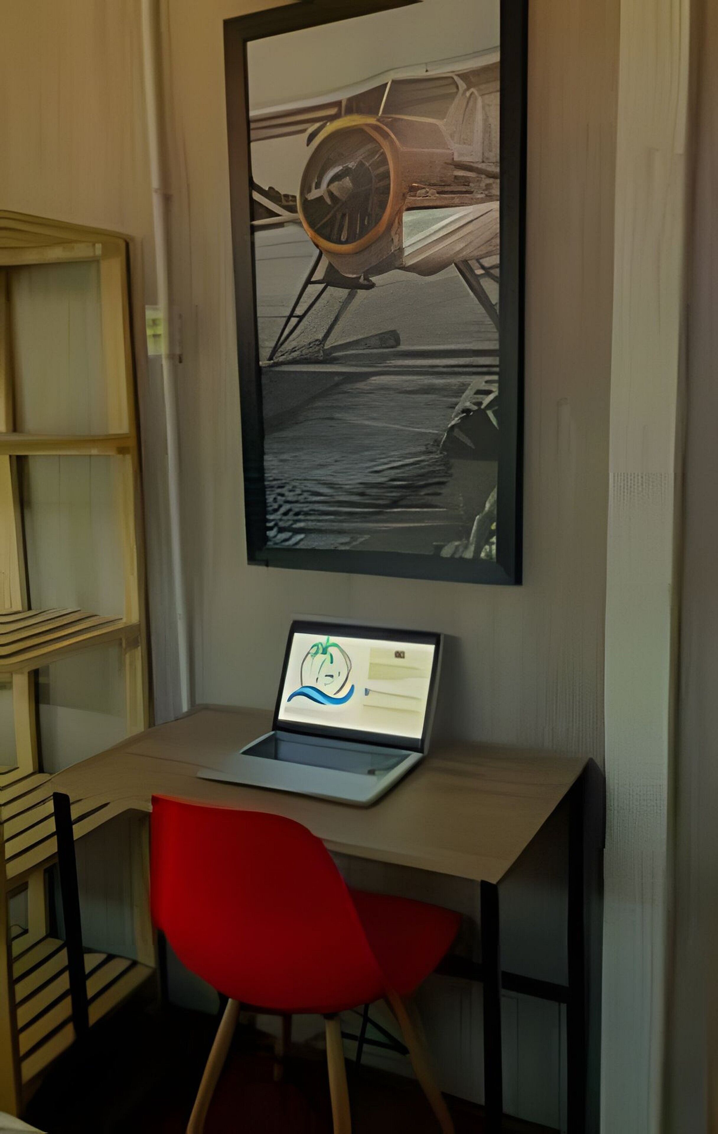 Et skrivebord med en bærbar computer, en rød stol og et billede i ramme af et skibsrat.