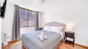 A neatly made bed with a grey bedspread and a rolled towel, a bedside table with a lamp, a wall-mounted TV, and a window with curtains. - Espacio Lira - 1D & Wifi (Santiago)