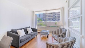 A modern living room with a grey sofa, striped armchair, and ottoman. Large windows offer a view of high-rise buildings. - Espacio Lira - 1D & Wifi (Santiago)