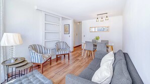 A modern living room with a grey sofa, striped armchairs, a small round table with a lamp, and a dining area with a wooden table and chairs. - Espacio Lira - 1D & Wifi (Santiago)