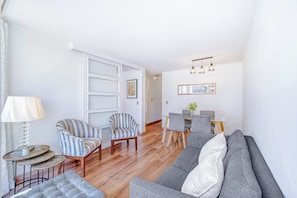 Ein modernes Wohnzimmer mit grauem Sofa, gestreiften Sesseln, einem kleinen runden Tisch mit Lampe sowie einem Essbereich mit Holztisch und Stühlen.