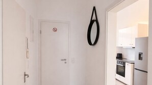 Un couloir étroit avec une porte blanche, un objet noir suspendu et une cuisine visible à travers une porte ouverte.