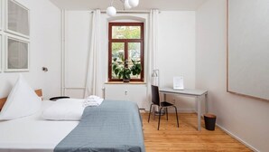 A hotel room with a bed, a desk, a chair, a window with curtains, a plant, and a wall-mounted light fixture. - Artist's room 1.5 - the minimalist (Gera)