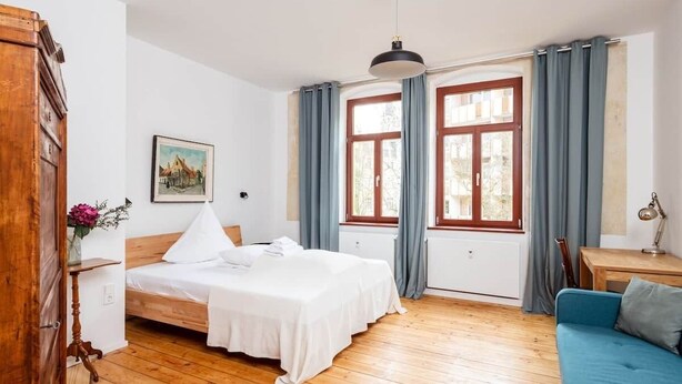 Une chambre avec un plancher en bois, un lit avec une literie blanche, un canapé bleu, une armoire en bois et une fenêtre avec des rideaux.