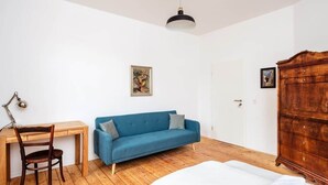 Une pièce avec un bureau en bois, un canapé bleu, une armoire en bois et un lit recouvert d’une couette blanche.