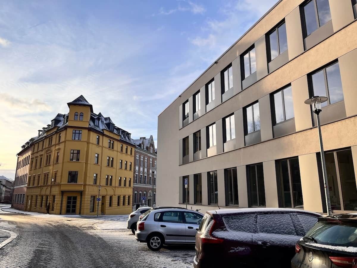 Une rue bordée de voitures garées et d’immeubles, dont un immeuble jaune et un immeuble moderne doté de grandes fenêtres.