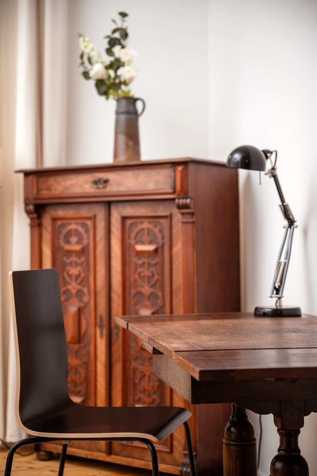 A wooden desk with a black desk lamp, a black chair, and a wooden cabinet with a vase of flowers.
