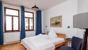 A hotel room with a wooden floor, a bed with white linens, a painting on the wall, and a desk with a lamp. - Artist room 2.2 - the spacious one (Gera)