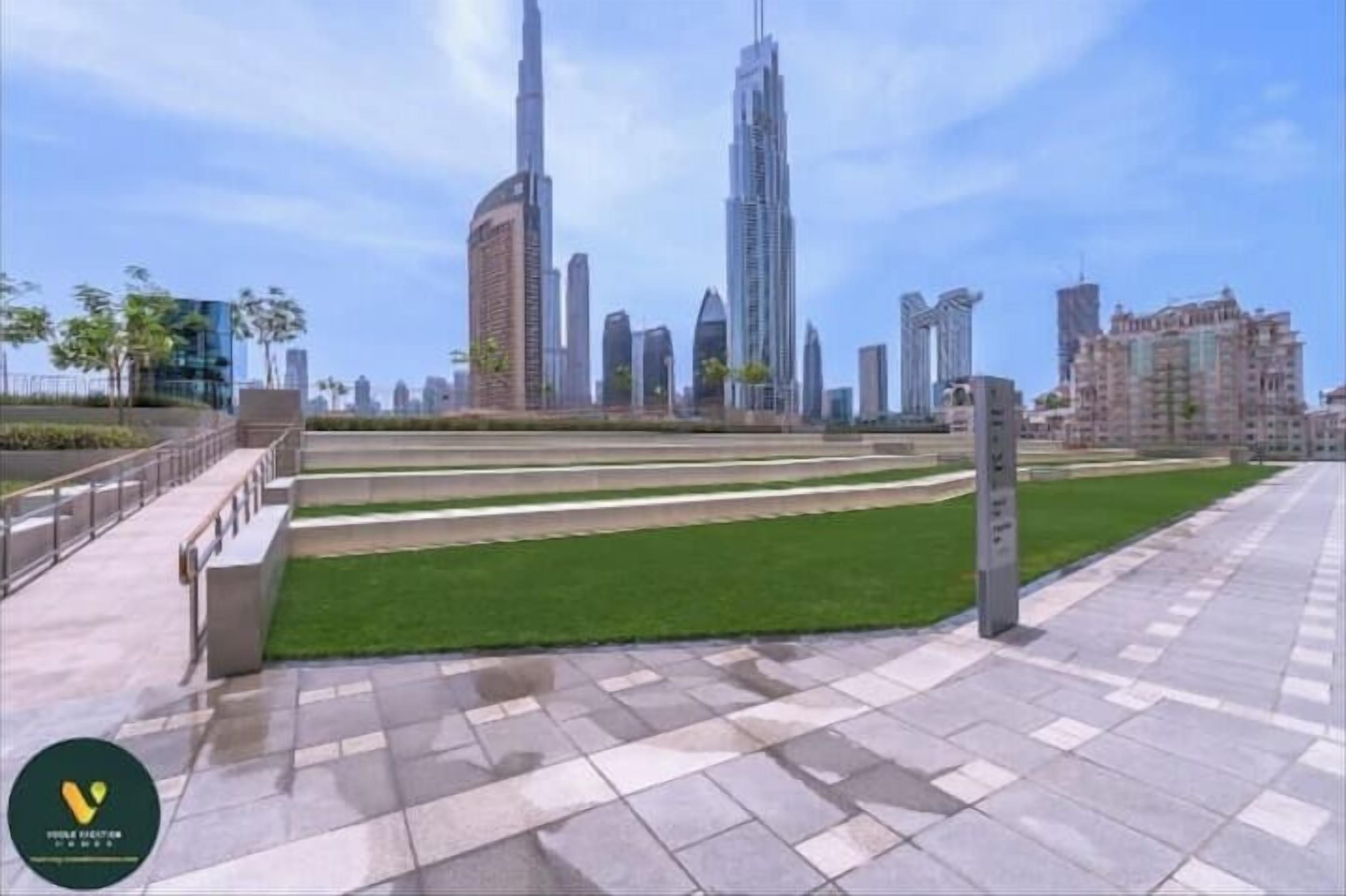 A cityscape with modern high-rise buildings, a central grassy area, and a paved walkway.