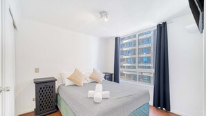 Un dormitorio con cama, mesita de noche, ventana con cortinas y vistas a edificios.