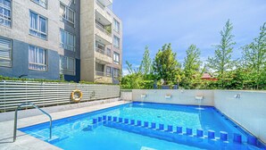 Una piscina con una escalera metálica, rodeada por una cerca blanca y contigua a un edificio de varios pisos con balcones.