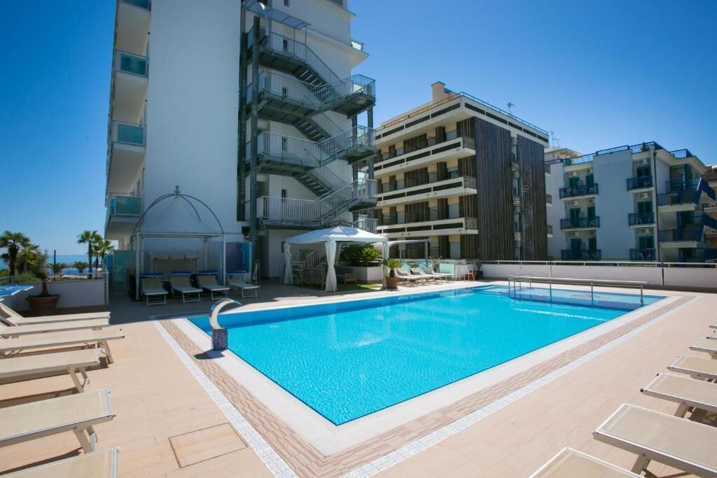 A rooftop swimming pool with lounge chairs, a covered seating area, and multi-story buildings in the background.