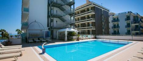 A rooftop swimming pool with lounge chairs, a covered seating area, and multi-story buildings in the background.