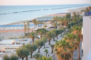 A beach with rows of umbrellas and palm trees, a view of the sea, and buildings in the background. - HOTEL  ALTIS  (San Benedetto del Tronto)