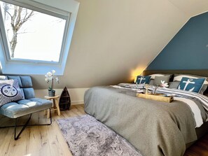 A bedroom with a large window, a bed with nautical-themed pillows, a small side table, and a bench. - Modern vacation home "Anker 2" with fireplace, underfloor heating & garden with dog (Trassenheide)