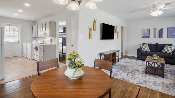 Ein modernes Wohnzimmer mit einem dunkelgrauen Sofa, einem Couchtisch aus Holz und einem Essbereich mit Holztisch und -stühlen. An der Wand ist ein Fernseher angebracht und es gibt einen Deckenventilator.
