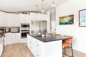 Une cuisine moderne avec des meubles blancs, un grand îlot central, des appareils en acier inoxydable et des luminaires suspendus.