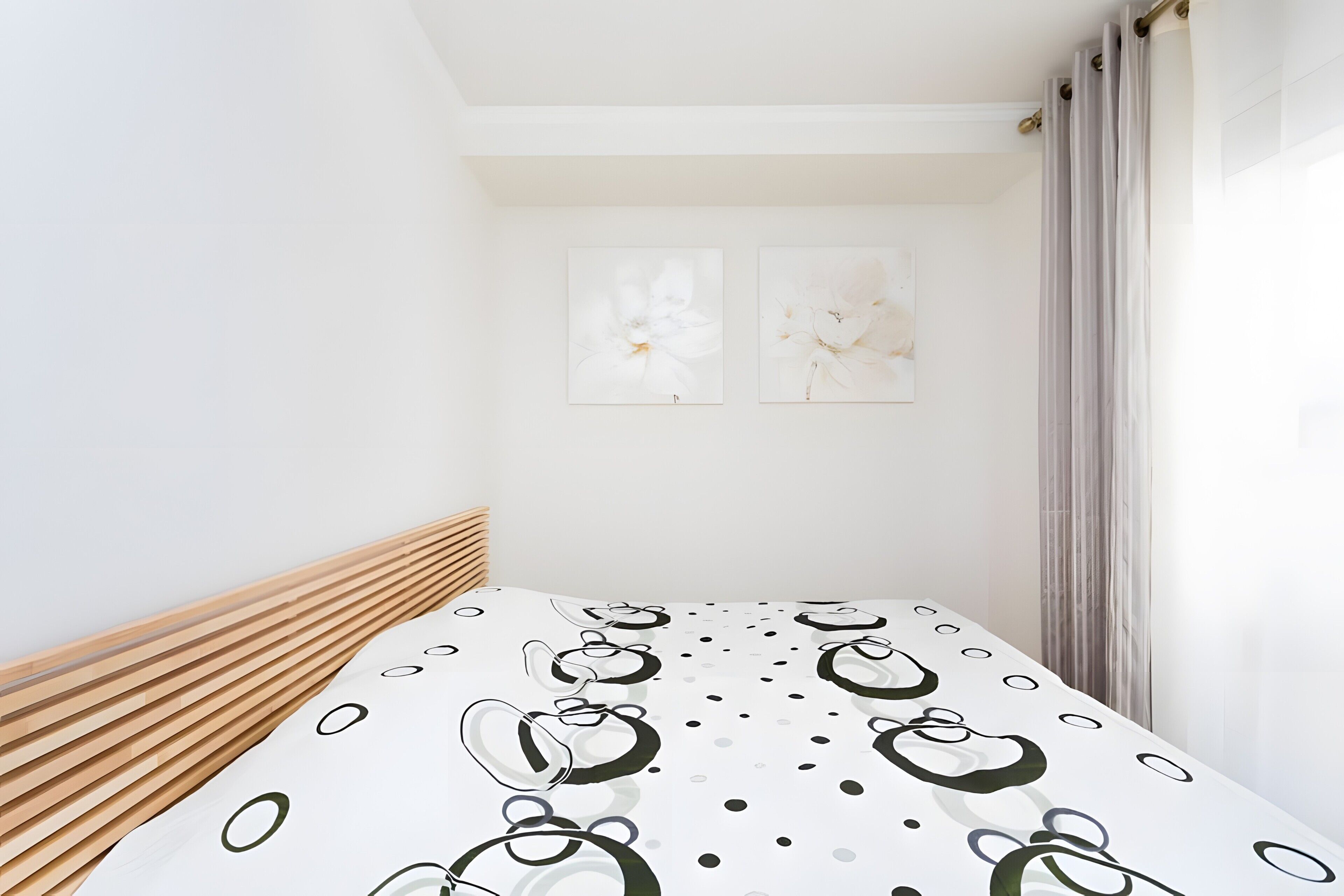 A bedroom with a bed, a wooden headboard, two wall-mounted paintings, and a window with curtains.