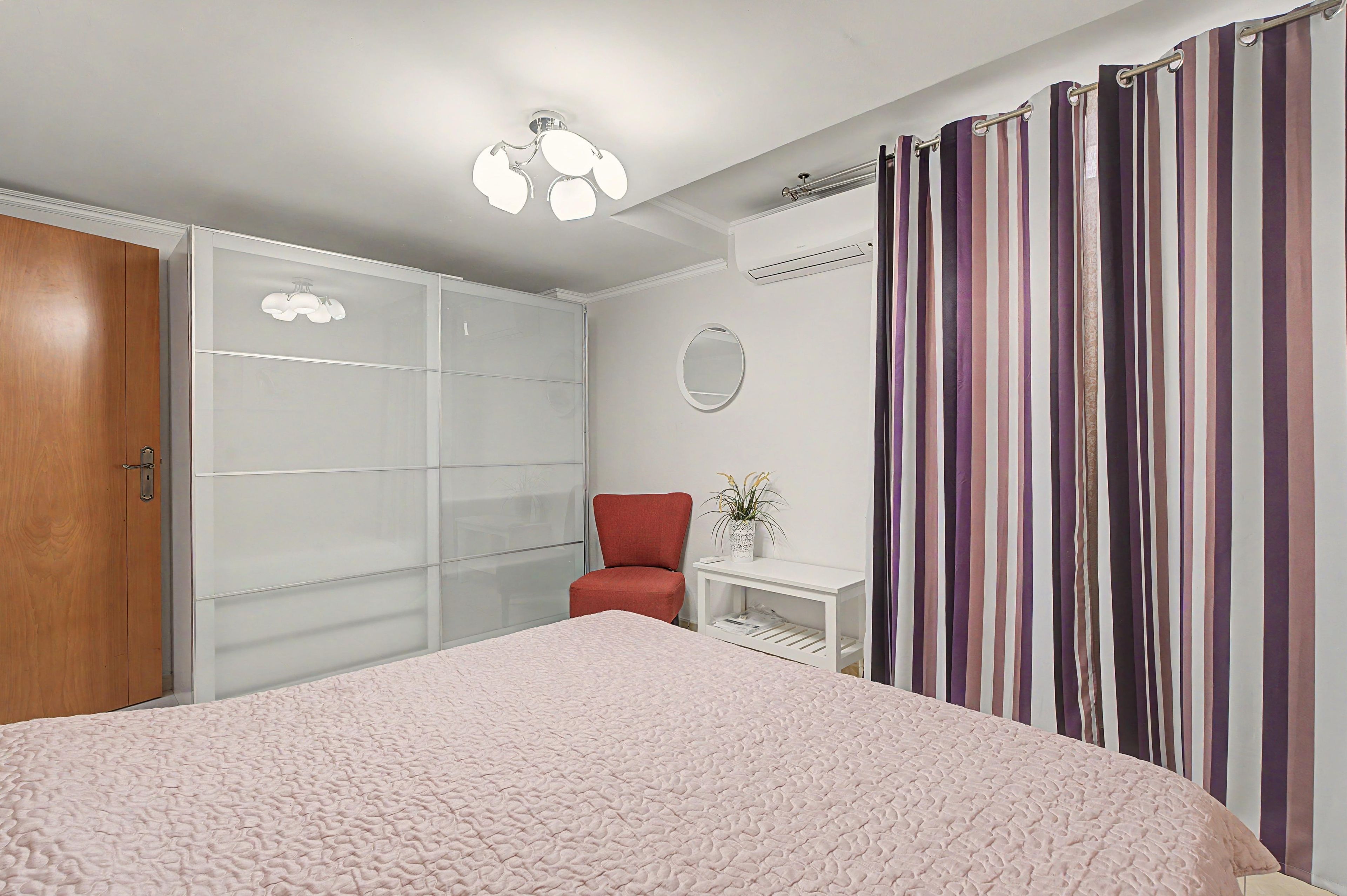 A bedroom with a bed, a wardrobe, a chair, a bedside table with a plant, and a curtain.
