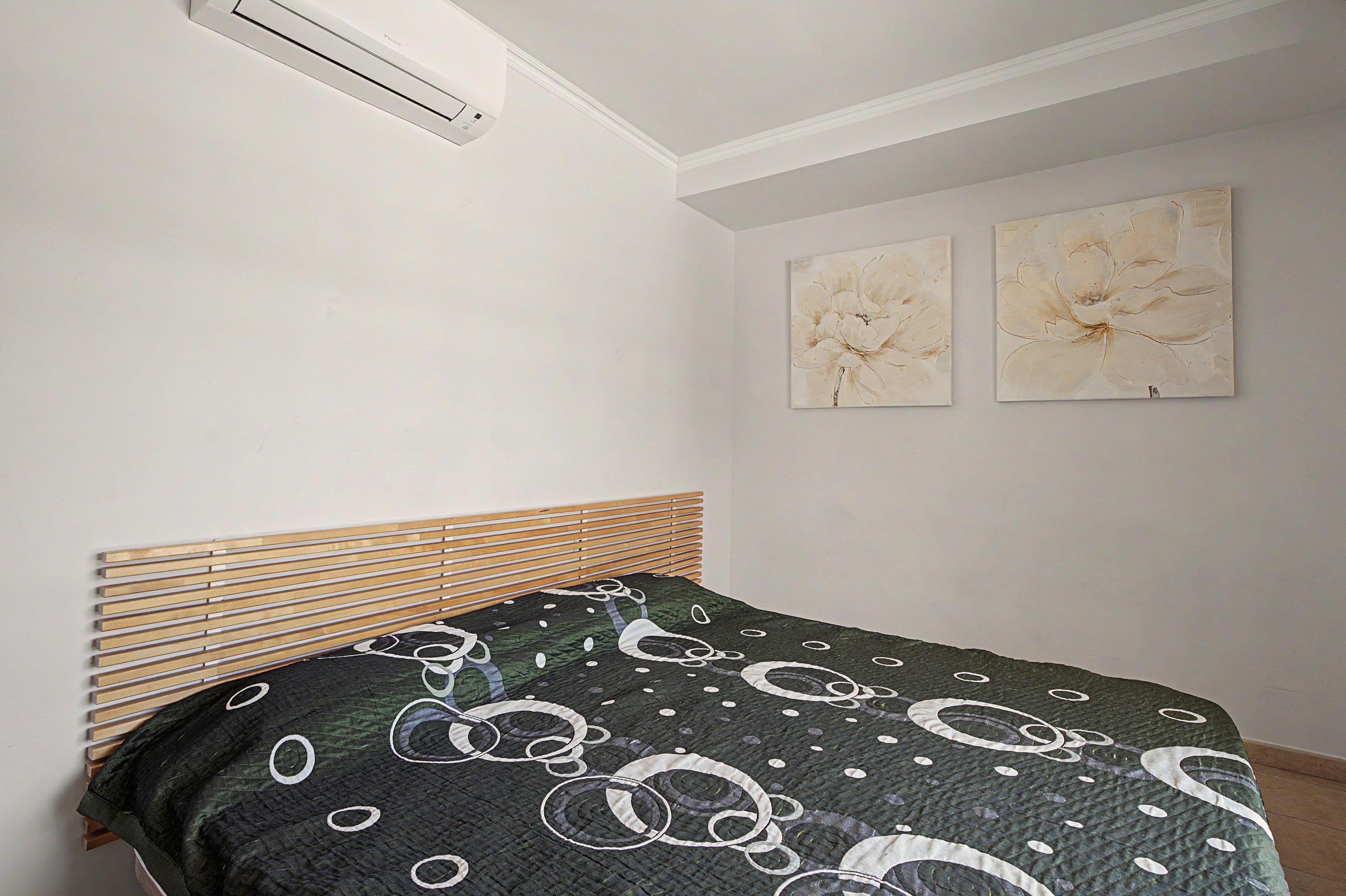 A bedroom with a bed, a headboard, two wall-mounted paintings, and a ceiling vent.