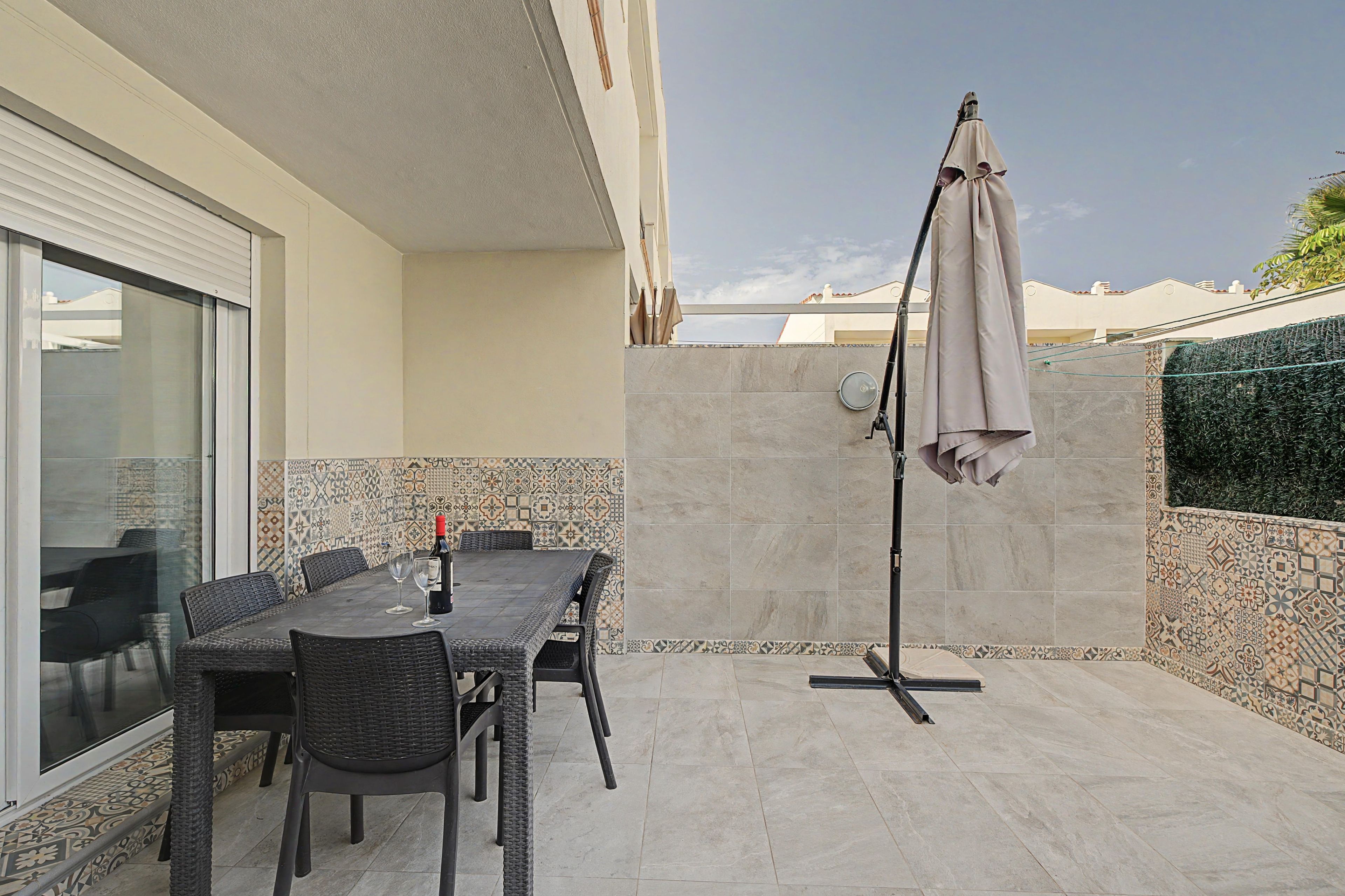 An outdoor patio with a table and chairs, a standing umbrella, and a tiled wall with a geometric pattern.