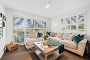 A spacious living room with a beige sectional sofa, a coffee table with a vase of flowers, and large windows with blinds. - Henley Horizon Apartment (Oxfordshire)