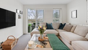 Een moderne woonkamer met een beige bank, een glazen salontafel, een groen vloerkleed en een grote tv die aan de muur is bevestigd.