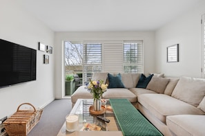 A modern living room with a beige sofa, a glass coffee table, a green rug, and a large TV mounted on the wall. - Henley Horizon Apartment (Oxfordshire)