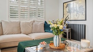 Een woonkamer met een beige bank, een vaas met bloemen, een fruitschaal, een kaars en een lijst met kunstwerk aan de muur.