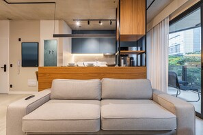 Un salon moderne avec un canapé, un coin cuisine doté d’un plan de travail en bois, et une vue sur l’extérieur.