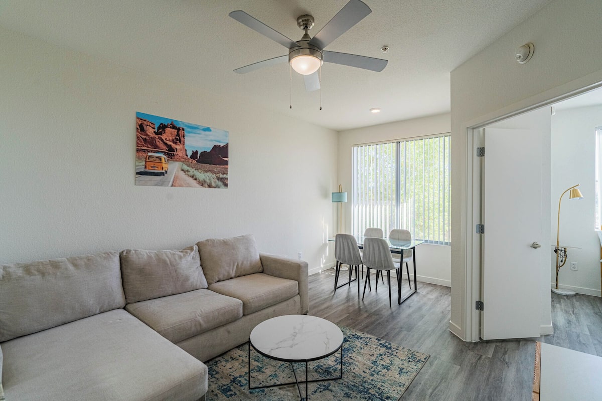 A living room with a beige sofa, a small round coffee table, a ceiling fan, and a painting of a desert scene.
