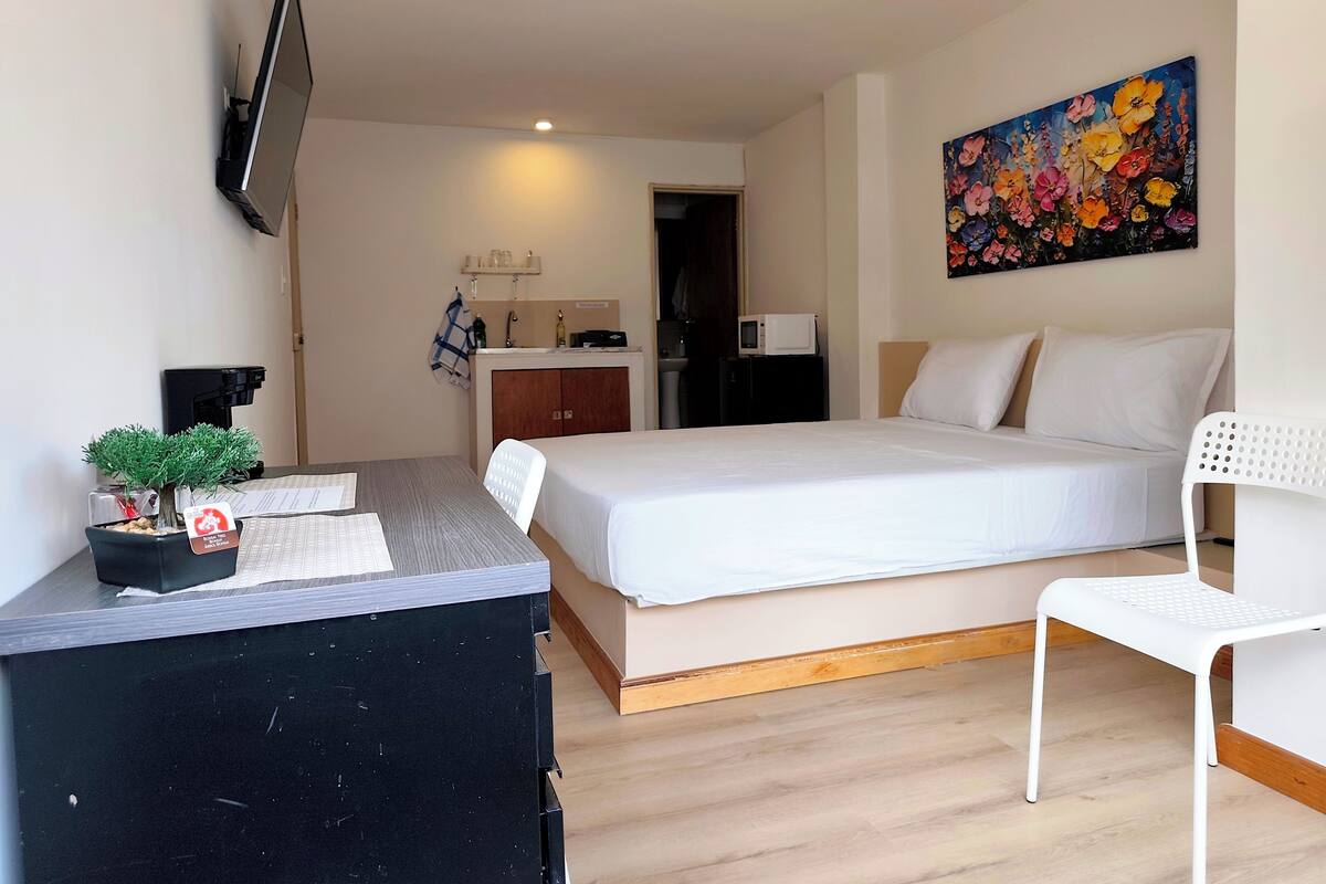 Une chambre d’hôtel avec un lit, un bureau agrémenté d’une plante, un téléviseur fixé au mur, un four à micro-ondes et un tableau représentant des fleurs.