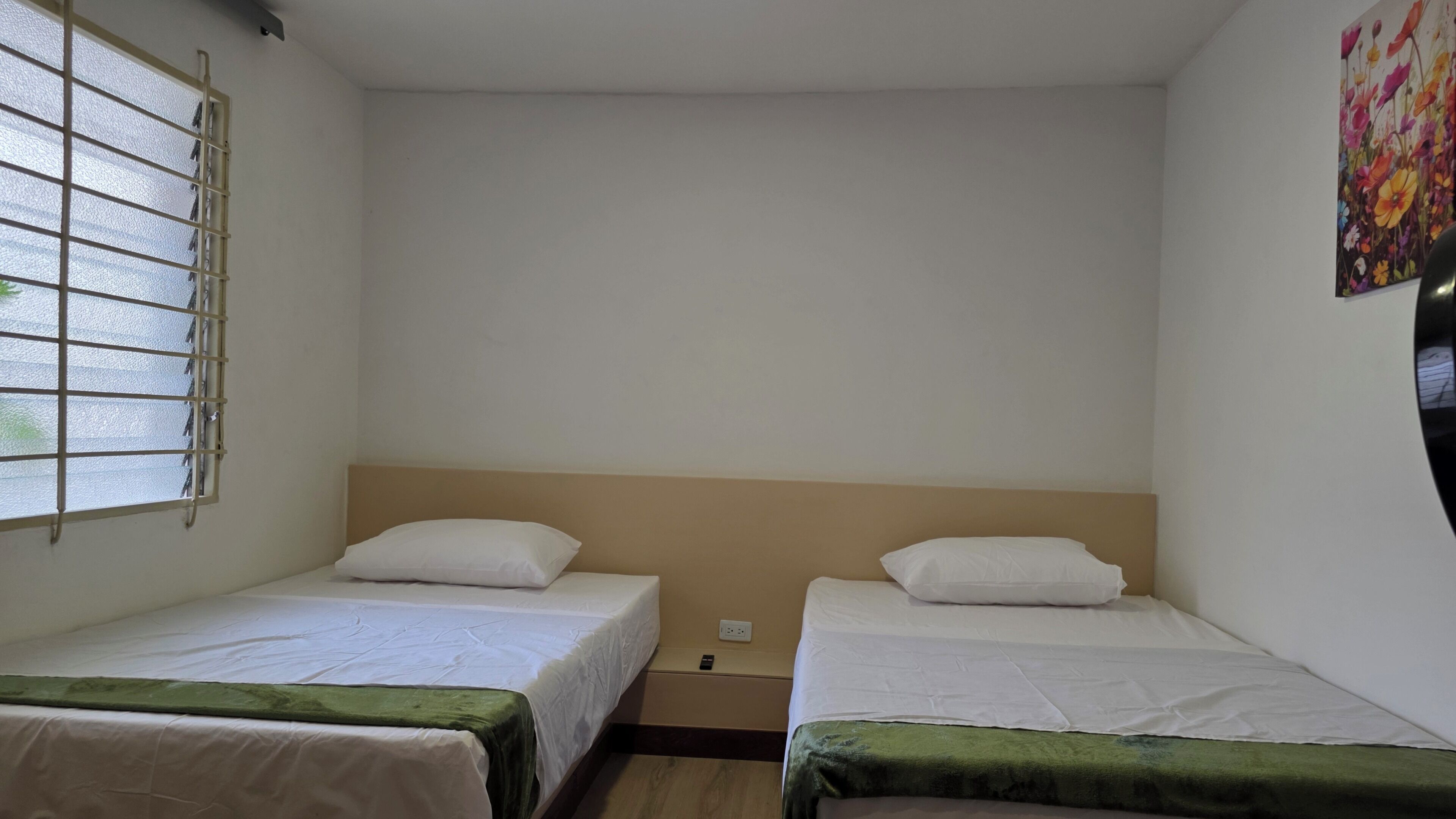 A small, clean room with two single beds, a window with bars, and a plain wall with a colorful painting.