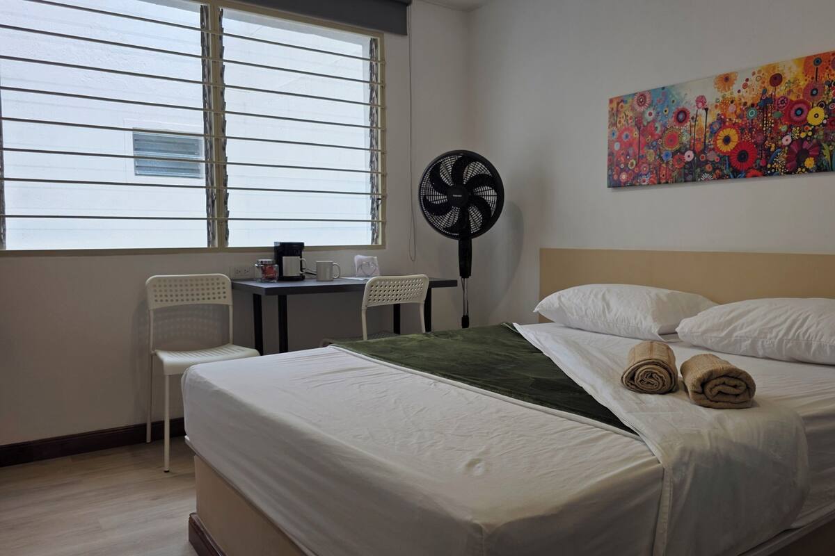 Une chambre d’hôtel avec un lit, un bureau, deux chaises, un ventilateur et un tableau coloré au mur.