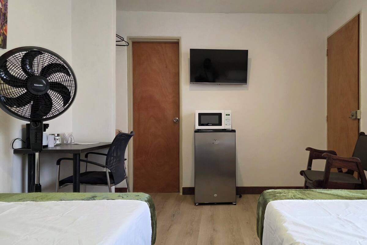 Une petite chambre d’hôtel avec deux lits, un bureau avec une chaise, un réfrigérateur, un four à micro-ondes, une télévision et un ventilateur.