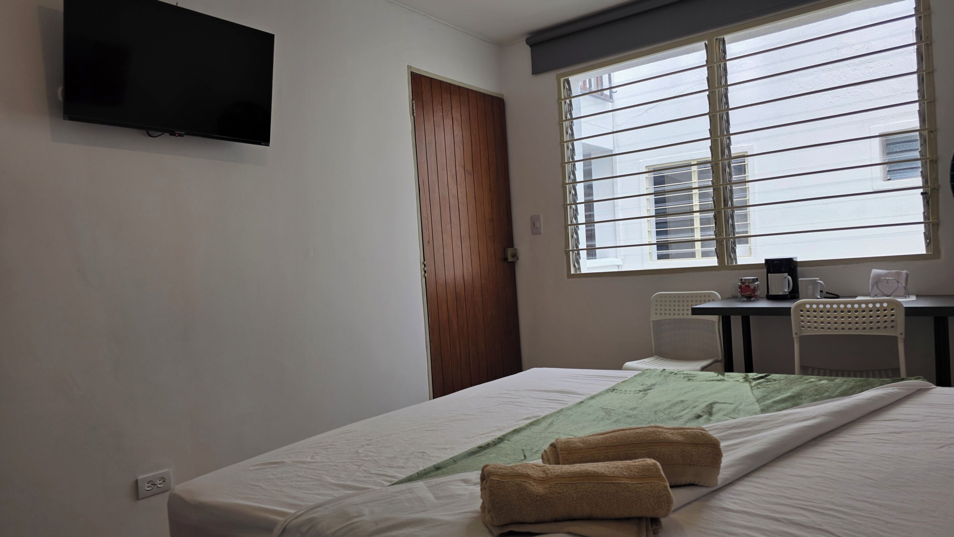 Habitación de hotel con cama, escritorio con silla, televisor montado en la pared y ventana con persianas.