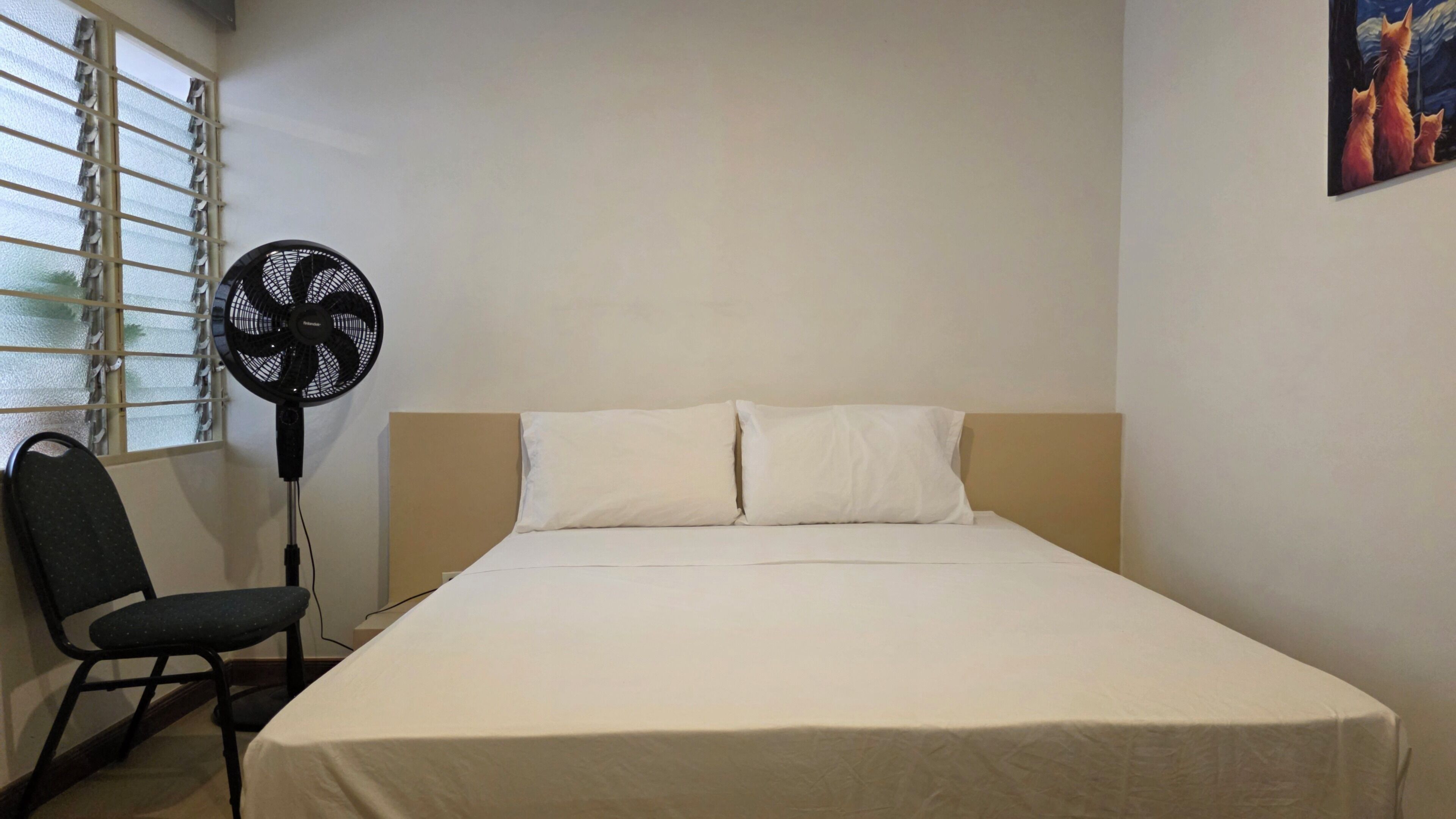 A bedroom with a bed, a chair, a standing fan, and a window with blinds.