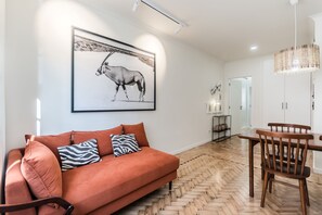 A modern living room with a herringbone wooden floor, a red sofa with patterned cushions, a dining table with wooden chairs, and a framed black and white photograph on the wall. - Casas da Tapada 1 In front of the wonderful Tapada das Necessidades (Lisboa)