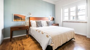 Ein Schlafzimmer mit einem Bett, Nachttischen aus Holz, einer Nachttischlampe, einem Wandspiegel und einem Fenster mit Jalousien.