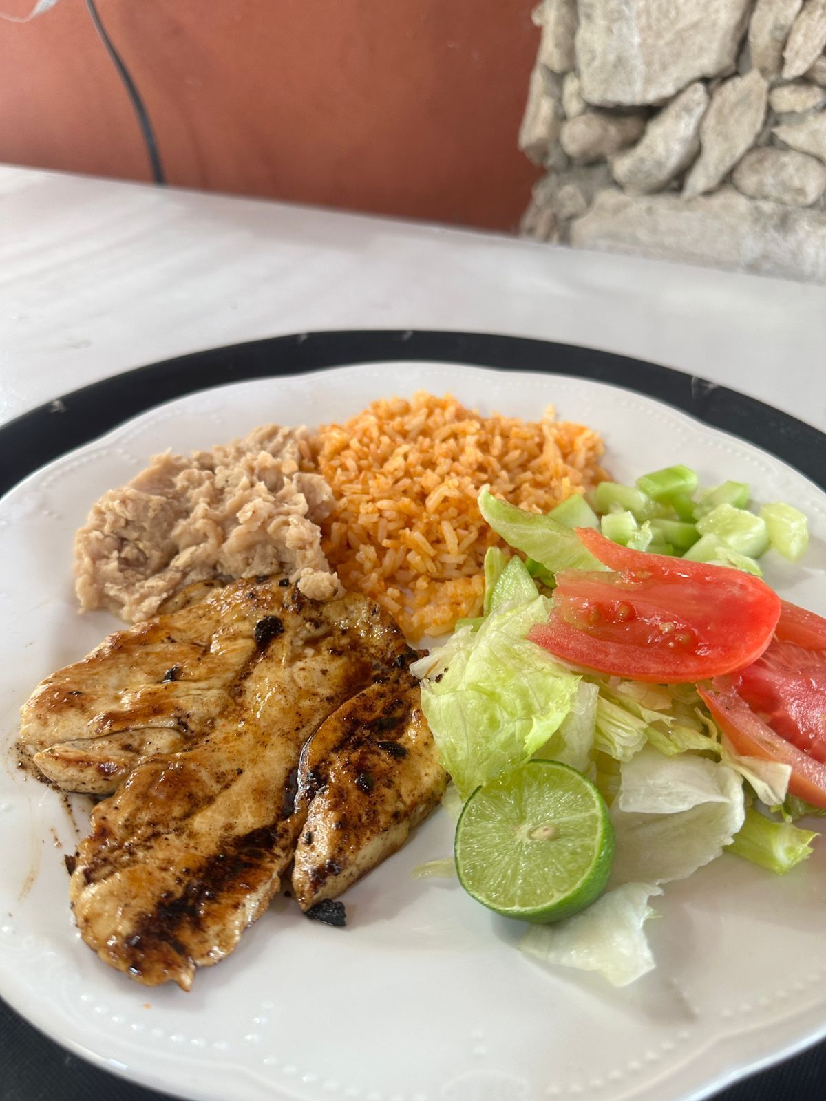 A plate with grilled chicken, rice, beans, lettuce, tomato, and lime.