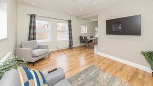 A modern living room with a grey sofa, a coffee table, and a flat-screen TV mounted on the wall. There are two windows with curtains, a dining area with a table and chairs, and a wooden floor. - Duplex Apartment - Modern & Convenient - Pass the Keys (Reading)