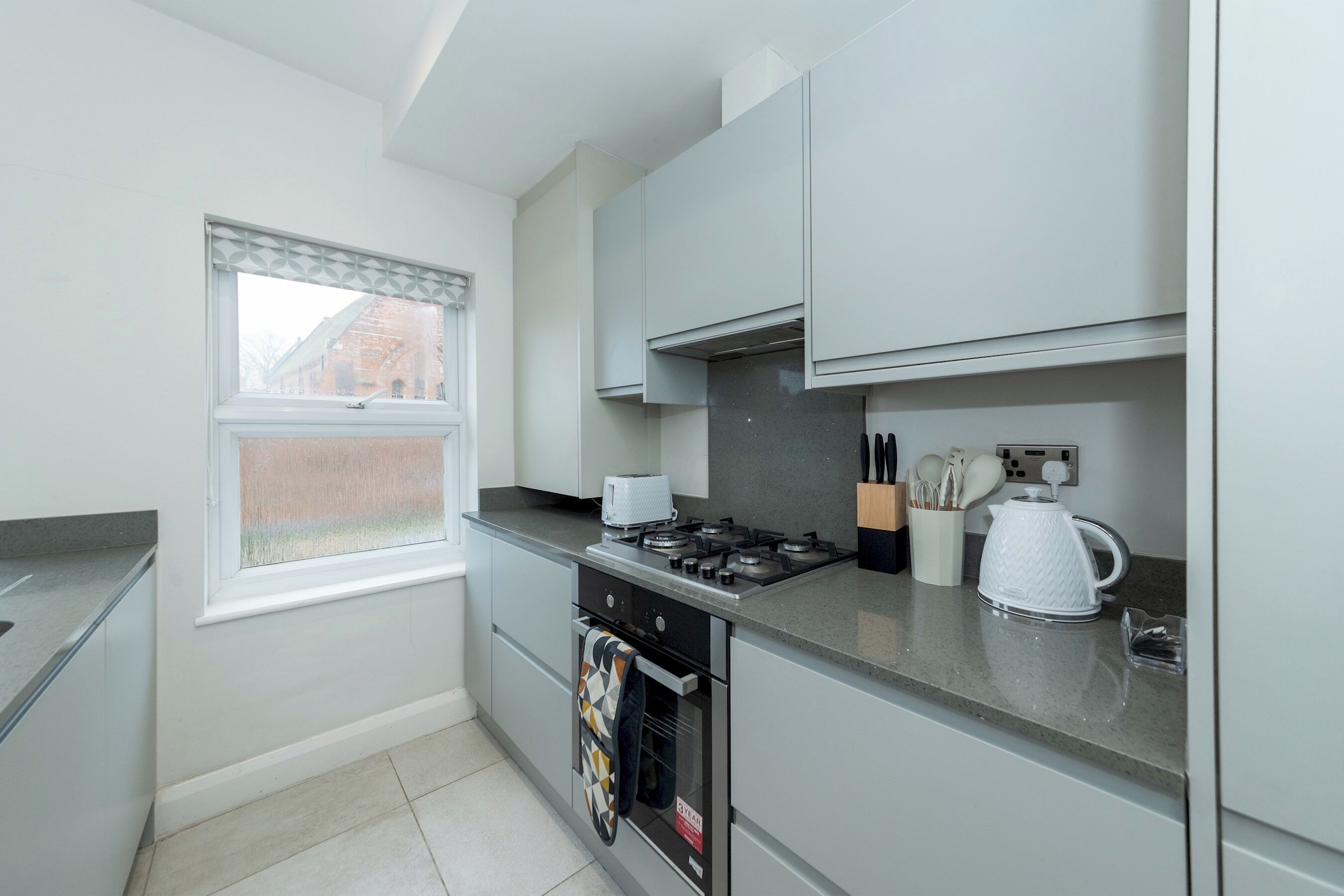 A modern kitchen with grey cabinets, a gas stove, a kettle, and a window with blinds.