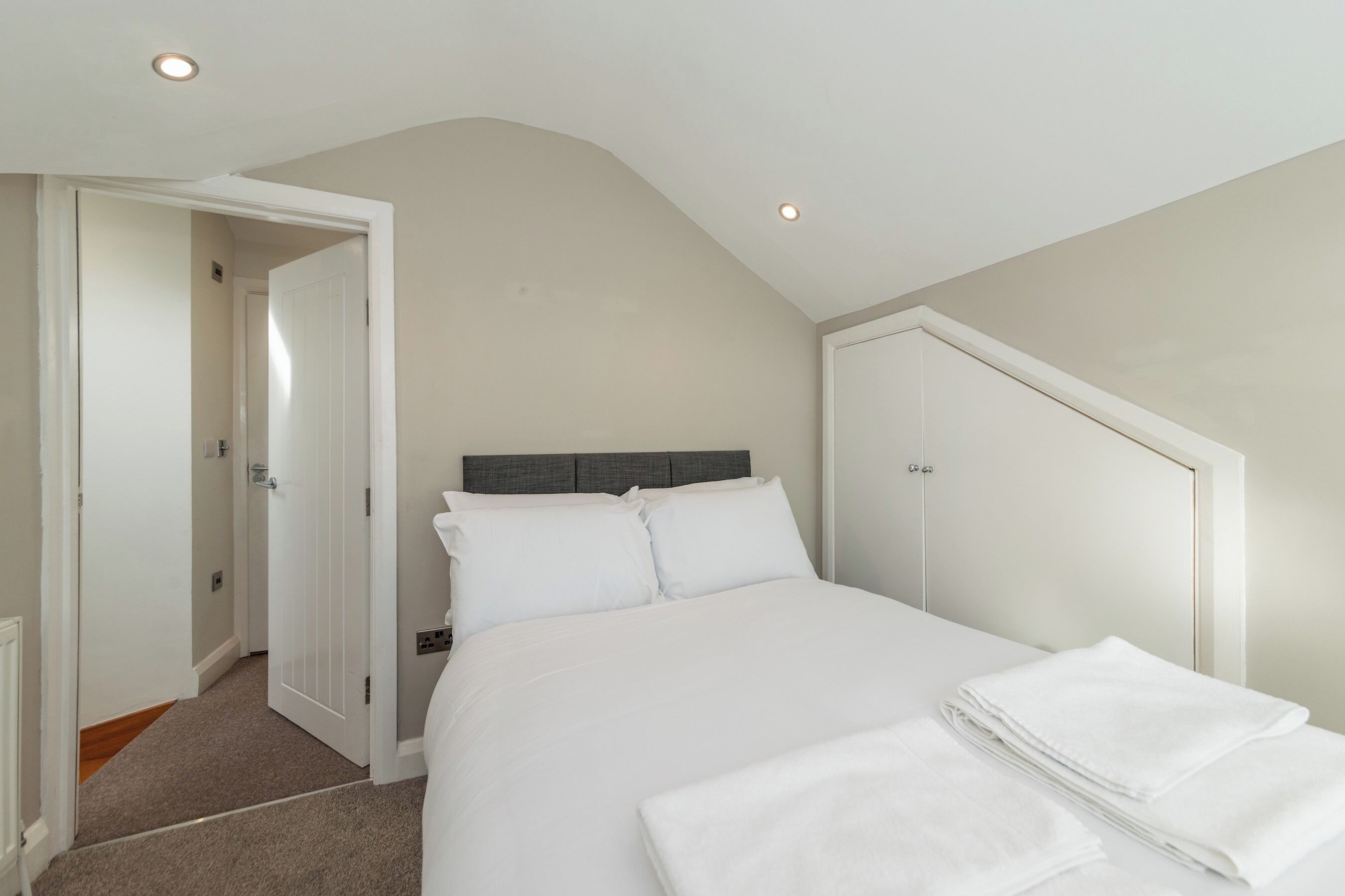 Een slaapkamer met een bed, witte lakens, een grijs hoofdeinde en een witte kast.