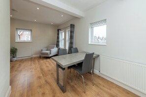A modern dining area with a wooden floor, a grey table, and black chairs. There are two windows with curtains, a radiator, and a potted plant. - Duplex Apartment - Modern & Convenient - Pass the Keys (Reading)