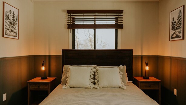Ein Schlafzimmer mit einem großen Bett, zwei Nachttischen mit Lampen, einem Fenster mit Jalousien und gerahmten Bildern an den Wänden.