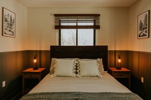 Une chambre à coucher avec un grand lit, deux tables de chevet avec des lampes, une fenêtre avec des stores et des tableaux encadrés accrochés aux murs.