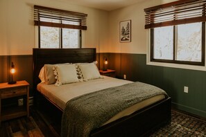 Una cama bien hecha con cabecera de madera, flanqueada por dos mesitas de noche con lámparas, en una habitación con paredes de paneles de madera y ventanas amplias con persianas.