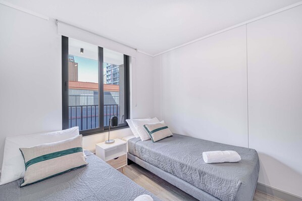 A modern bedroom with two beds, a nightstand, a window with a view of buildings, and a wardrobe. - Urban Essence - 2D Santiago (Santiago)