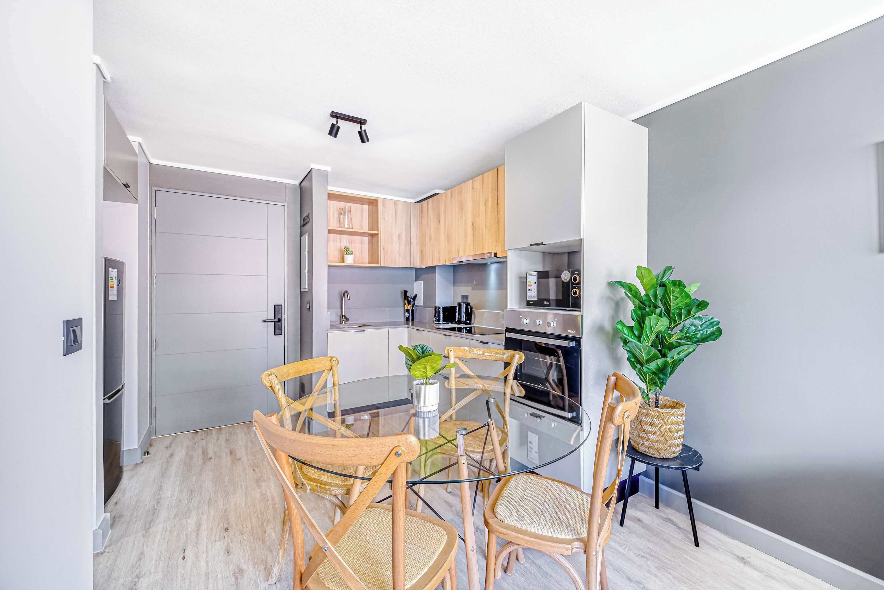 A modern kitchen with wooden cabinets, a glass dining table, and chairs. There is a potted plant on the side.